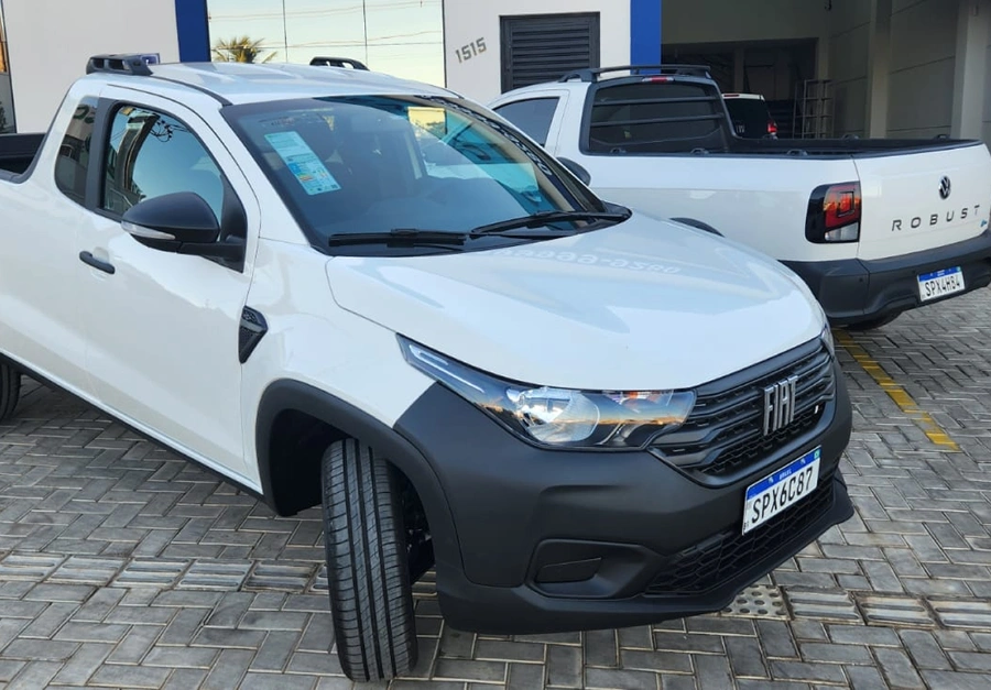 Picape Fiat Strada branca estacionada em frente à UP Locadora de Veículos, com outra picape Volkswagen Robust ao fundo.