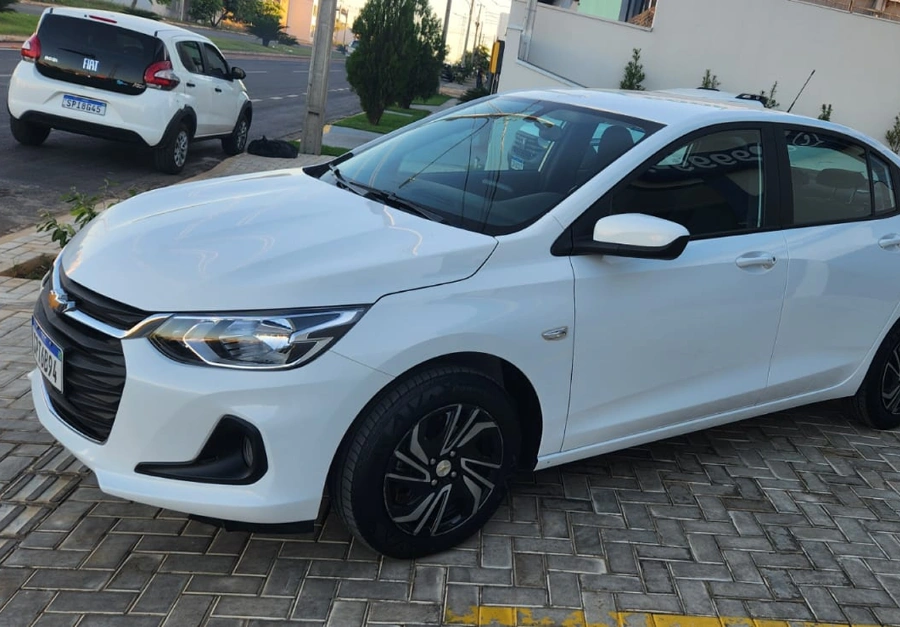 Chevrolet sedan branco da frota da UP Locadora de Veículos estacionado na entrada, com um Fiat Mobi ao fundo.